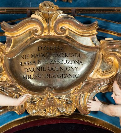 Zdjęcie nr 24: Ołtarz architektoniczny, jednoosiowy, jednokondygnacyjny ze zwieńczeniem. Na zdwojonych cokołach usytuowanych ukośnie względem osi po trzy kompozytowe kolumny opinające filary o gładkich trzonach podtrzymujących wyłamujące się nad nimi belkowanie z ćwierćkolistymi naczółkami. Na nich siedzące anioły z narzędzami męki Pańskiej. Pośrodku wnęka zamknięta półkoliście i wchodząca w partię belkowania z figurą Ukrzyżowanego. Nad wnęką dwa nagie putta trzymają kartusz z napisem: &quot;DZIEŁO / NIE MAJĄC PRZYKŁADU / ŁASKA NIE ZASŁUŻONA / DAR NIE OCENIONY / MIŁOŚĆ BEZ GRANIC&quot;.  Zwieńczenie ujęte spływami wolutowymi, mocno zwężające się ku górze, zakończone gzymsem w formie łuku nadwieszonego. W polu owalny prześwit z glorią promienistą w chmurach z figurą Boga Ojca, puttami siedzącymi na gzymsie i główkami aniołów wśród chmur. Po bokach ołtarza dwie bramki z cokołami ujętymi spływami wolutowymi, na których figury św. Jana Chrzciciela i św. Jana Ewangelisty. Dekoracja ornamentalna w postaci rozłożystych liści na cokołach oraz liściastych, ażurowych uszy ujmujących nastawę. 