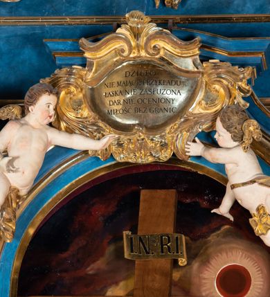 Zdjęcie nr 31: Ołtarz architektoniczny, jednoosiowy, jednokondygnacyjny ze zwieńczeniem. Na zdwojonych cokołach usytuowanych ukośnie względem osi po trzy kompozytowe kolumny opinające filary o gładkich trzonach podtrzymujących wyłamujące się nad nimi belkowanie z ćwierćkolistymi naczółkami. Na nich siedzące anioły z narzędzami męki Pańskiej. Pośrodku wnęka zamknięta półkoliście i wchodząca w partię belkowania z figurą Ukrzyżowanego. Nad wnęką dwa nagie putta trzymają kartusz z napisem: "DZIEŁO / NIE MAJĄC PRZYKŁADU / ŁASKA NIE ZASŁUŻONA / DAR NIE OCENIONY / MIŁOŚĆ BEZ GRANIC".  Zwieńczenie ujęte spływami wolutowymi, mocno zwężające się ku górze, zakończone gzymsem w formie łuku nadwieszonego. W polu owalny prześwit z glorią promienistą w chmurach z figurą Boga Ojca, puttami siedzącymi na gzymsie i główkami aniołów wśród chmur. Po bokach ołtarza dwie bramki z cokołami ujętymi spływami wolutowymi, na których figury św. Jana Chrzciciela i św. Jana Ewangelisty. Dekoracja ornamentalna w postaci rozłożystych liści na cokołach oraz liściastych, ażurowych uszy ujmujących nastawę. 