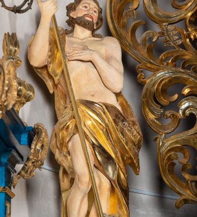 Zdjęcie nr 64: Ołtarz architektoniczny, jednoosiowy, jednokondygnacyjny ze zwieńczeniem. Na zdwojonych cokołach usytuowanych ukośnie względem osi po trzy kompozytowe kolumny opinające filary o gładkich trzonach podtrzymujących wyłamujące się nad nimi belkowanie z ćwierćkolistymi naczółkami. Na nich siedzące anioły z narzędzami męki Pańskiej. Pośrodku wnęka zamknięta półkoliście i wchodząca w partię belkowania z figurą Ukrzyżowanego. Nad wnęką dwa nagie putta trzymają kartusz z napisem: "DZIEŁO / NIE MAJĄC PRZYKŁADU / ŁASKA NIE ZASŁUŻONA / DAR NIE OCENIONY / MIŁOŚĆ BEZ GRANIC".  Zwieńczenie ujęte spływami wolutowymi, mocno zwężające się ku górze, zakończone gzymsem w formie łuku nadwieszonego. W polu owalny prześwit z glorią promienistą w chmurach z figurą Boga Ojca, puttami siedzącymi na gzymsie i główkami aniołów wśród chmur. Po bokach ołtarza dwie bramki z cokołami ujętymi spływami wolutowymi, na których figury św. Jana Chrzciciela i św. Jana Ewangelisty. Dekoracja ornamentalna w postaci rozłożystych liści na cokołach oraz liściastych, ażurowych uszy ujmujących nastawę. 