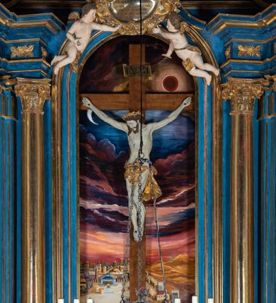 Zdjęcie nr 87: Ołtarz architektoniczny, jednoosiowy, jednokondygnacyjny ze zwieńczeniem. Na zdwojonych cokołach usytuowanych ukośnie względem osi po trzy kompozytowe kolumny opinające filary o gładkich trzonach podtrzymujących wyłamujące się nad nimi belkowanie z ćwierćkolistymi naczółkami. Na nich siedzące anioły z narzędzami męki Pańskiej. Pośrodku wnęka zamknięta półkoliście i wchodząca w partię belkowania z figurą Ukrzyżowanego. Nad wnęką dwa nagie putta trzymają kartusz z napisem: &quot;DZIEŁO / NIE MAJĄC PRZYKŁADU / ŁASKA NIE ZASŁUŻONA / DAR NIE OCENIONY / MIŁOŚĆ BEZ GRANIC&quot;.  Zwieńczenie ujęte spływami wolutowymi, mocno zwężające się ku górze, zakończone gzymsem w formie łuku nadwieszonego. W polu owalny prześwit z glorią promienistą w chmurach z figurą Boga Ojca, puttami siedzącymi na gzymsie i główkami aniołów wśród chmur. Po bokach ołtarza dwie bramki z cokołami ujętymi spływami wolutowymi, na których figury św. Jana Chrzciciela i św. Jana Ewangelisty. Dekoracja ornamentalna w postaci rozłożystych liści na cokołach oraz liściastych, ażurowych uszy ujmujących nastawę. 