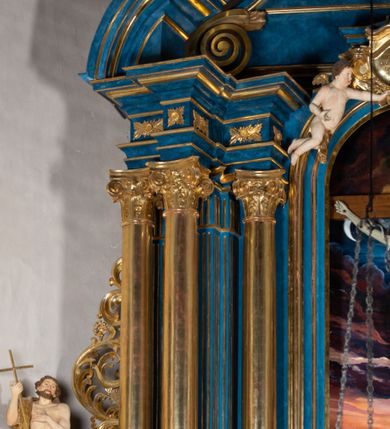 Zdjęcie nr 90: Ołtarz architektoniczny, jednoosiowy, jednokondygnacyjny ze zwieńczeniem. Na zdwojonych cokołach usytuowanych ukośnie względem osi po trzy kompozytowe kolumny opinające filary o gładkich trzonach podtrzymujących wyłamujące się nad nimi belkowanie z ćwierćkolistymi naczółkami. Na nich siedzące anioły z narzędzami męki Pańskiej. Pośrodku wnęka zamknięta półkoliście i wchodząca w partię belkowania z figurą Ukrzyżowanego. Nad wnęką dwa nagie putta trzymają kartusz z napisem: "DZIEŁO / NIE MAJĄC PRZYKŁADU / ŁASKA NIE ZASŁUŻONA / DAR NIE OCENIONY / MIŁOŚĆ BEZ GRANIC".  Zwieńczenie ujęte spływami wolutowymi, mocno zwężające się ku górze, zakończone gzymsem w formie łuku nadwieszonego. W polu owalny prześwit z glorią promienistą w chmurach z figurą Boga Ojca, puttami siedzącymi na gzymsie i główkami aniołów wśród chmur. Po bokach ołtarza dwie bramki z cokołami ujętymi spływami wolutowymi, na których figury św. Jana Chrzciciela i św. Jana Ewangelisty. Dekoracja ornamentalna w postaci rozłożystych liści na cokołach oraz liściastych, ażurowych uszy ujmujących nastawę. 