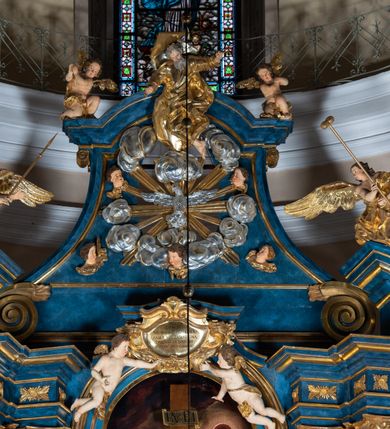 Zdjęcie nr 91: Ołtarz architektoniczny, jednoosiowy, jednokondygnacyjny ze zwieńczeniem. Na zdwojonych cokołach usytuowanych ukośnie względem osi po trzy kompozytowe kolumny opinające filary o gładkich trzonach podtrzymujących wyłamujące się nad nimi belkowanie z ćwierćkolistymi naczółkami. Na nich siedzące anioły z narzędzami męki Pańskiej. Pośrodku wnęka zamknięta półkoliście i wchodząca w partię belkowania z figurą Ukrzyżowanego. Nad wnęką dwa nagie putta trzymają kartusz z napisem: "DZIEŁO / NIE MAJĄC PRZYKŁADU / ŁASKA NIE ZASŁUŻONA / DAR NIE OCENIONY / MIŁOŚĆ BEZ GRANIC".  Zwieńczenie ujęte spływami wolutowymi, mocno zwężające się ku górze, zakończone gzymsem w formie łuku nadwieszonego. W polu owalny prześwit z glorią promienistą w chmurach z figurą Boga Ojca, puttami siedzącymi na gzymsie i główkami aniołów wśród chmur. Po bokach ołtarza dwie bramki z cokołami ujętymi spływami wolutowymi, na których figury św. Jana Chrzciciela i św. Jana Ewangelisty. Dekoracja ornamentalna w postaci rozłożystych liści na cokołach oraz liściastych, ażurowych uszy ujmujących nastawę. 