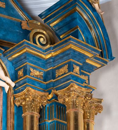 Zdjęcie nr 98: Ołtarz architektoniczny, jednoosiowy, jednokondygnacyjny ze zwieńczeniem. Na zdwojonych cokołach usytuowanych ukośnie względem osi po trzy kompozytowe kolumny opinające filary o gładkich trzonach podtrzymujących wyłamujące się nad nimi belkowanie z ćwierćkolistymi naczółkami. Na nich siedzące anioły z narzędzami męki Pańskiej. Pośrodku wnęka zamknięta półkoliście i wchodząca w partię belkowania z figurą Ukrzyżowanego. Nad wnęką dwa nagie putta trzymają kartusz z napisem: "DZIEŁO / NIE MAJĄC PRZYKŁADU / ŁASKA NIE ZASŁUŻONA / DAR NIE OCENIONY / MIŁOŚĆ BEZ GRANIC".  Zwieńczenie ujęte spływami wolutowymi, mocno zwężające się ku górze, zakończone gzymsem w formie łuku nadwieszonego. W polu owalny prześwit z glorią promienistą w chmurach z figurą Boga Ojca, puttami siedzącymi na gzymsie i główkami aniołów wśród chmur. Po bokach ołtarza dwie bramki z cokołami ujętymi spływami wolutowymi, na których figury św. Jana Chrzciciela i św. Jana Ewangelisty. Dekoracja ornamentalna w postaci rozłożystych liści na cokołach oraz liściastych, ażurowych uszy ujmujących nastawę. 