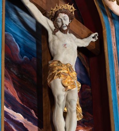 Zdjęcie nr 44: Ołtarz architektoniczny, jednoosiowy, jednokondygnacyjny ze zwieńczeniem. Na zdwojonych cokołach usytuowanych ukośnie względem osi po trzy kompozytowe kolumny opinające filary o gładkich trzonach podtrzymujących wyłamujące się nad nimi belkowanie z ćwierćkolistymi naczółkami. Na nich siedzące anioły z narzędzami męki Pańskiej. Pośrodku wnęka zamknięta półkoliście i wchodząca w partię belkowania z figurą Ukrzyżowanego. Nad wnęką dwa nagie putta trzymają kartusz z napisem: "DZIEŁO / NIE MAJĄC PRZYKŁADU / ŁASKA NIE ZASŁUŻONA / DAR NIE OCENIONY / MIŁOŚĆ BEZ GRANIC".  Zwieńczenie ujęte spływami wolutowymi, mocno zwężające się ku górze, zakończone gzymsem w formie łuku nadwieszonego. W polu owalny prześwit z glorią promienistą w chmurach z figurą Boga Ojca, puttami siedzącymi na gzymsie i główkami aniołów wśród chmur. Po bokach ołtarza dwie bramki z cokołami ujętymi spływami wolutowymi, na których figury św. Jana Chrzciciela i św. Jana Ewangelisty. Dekoracja ornamentalna w postaci rozłożystych liści na cokołach oraz liściastych, ażurowych uszy ujmujących nastawę. 