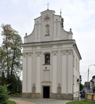 Zdjęcie nr 1:  Kościół usytuowany jest za północną pierzeją zabudowy rynku, w bezpośrednim sąsiedztwie świątyni parafialnej, po jej południowo-wschodniej stronie. 
Kościół orientowany, jest budowlą murowaną z cegły i kamienia (grubość murów od 70 do 160 cm), na rzucie podłużnym (24 m długość, 8,5 m szerokość). Korpus jednonawowy, trójprzęsłowy, prezbiterium jednoprzęsłowe, zamknięte półkolistą absydą, z dwukondygnacyjną przyległością od północy (zakrystia w przyziemiu). Nawę i prezbiterium przekrywa sklepienie kolebkowe z gurtami wspartymi na parach pilastrów dźwigających wydatne, obiegające wnętrze belkowanie. Nawę od prezbiterium oddziela półkolista arkada łuku tęczowego. Wnętrze doświetlają dwa rzędy okien zamkniętych łukami odcinkowymi. Artykulację ścian na zewnątrz stanowią płyciny ujmujące pary okien. Trójosiową fasadę artykułują pary osadzonych na cokole pilastrów – po dwie po bokach i jedna w polu środkowym wspierające belkowanie. W osi środkowej jest umiejscowiony ujęty profilowanym obramieniem otwór wejściowy z dwuskrzydłowymi drzwiami. Wyżej jest płytka, prostokątna wnęka, u góry zaś okno zamknięte łukiem odcinkowym. W osiach bocznych na cokole fasady osadzona para niszy z rzeźbami św. Piotra po lewej stronie i św. Pawła po prawej. Kapitele pilastrów zdobione motywami festonów owocowo-kwiatowych.   
Szczyt fasady ujęty parą spływów, zamknięty trójkątnie, po bokach flankowany parą cokołów zwieńczonych wazonami, pole środkowe wydzielone dwiema parami pilastrów zawiera półkoliście zamkniętą niszę z figurą Matki Boskiej, wyżej trójkąt Opatrzności Bożej ujęty promienistą glorią. Dachy korpusu i prezbiterium dwuspadowe, nad pierwszym przęsłem wieżyczka na sygnaturkę w formie latarni. Dach nad przyległością pulpitowy. Tynki kościoła w odcieniach bieli i szarości. 
