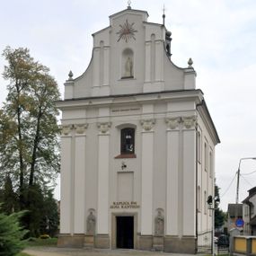 Zdjęcie nr 1:  Kościół usytuowany jest za północną pierzeją zabudowy rynku, w bezpośrednim sąsiedztwie świątyni parafialnej, po jej południowo-wschodniej stronie. 
Kościół orientowany, jest budowlą murowaną z cegły i kamienia (grubość murów od 70 do 160 cm), na rzucie podłużnym (24 m długość, 8,5 m szerokość). Korpus jednonawowy, trójprzęsłowy, prezbiterium jednoprzęsłowe, zamknięte półkolistą absydą, z dwukondygnacyjną przyległością od północy (zakrystia w przyziemiu). Nawę i prezbiterium przekrywa sklepienie kolebkowe z gurtami wspartymi na parach pilastrów dźwigających wydatne, obiegające wnętrze belkowanie. Nawę od prezbiterium oddziela półkolista arkada łuku tęczowego. Wnętrze doświetlają dwa rzędy okien zamkniętych łukami odcinkowymi. Artykulację ścian na zewnątrz stanowią płyciny ujmujące pary okien. Trójosiową fasadę artykułują pary osadzonych na cokole pilastrów – po dwie po bokach i jedna w polu środkowym wspierające belkowanie. W osi środkowej jest umiejscowiony ujęty profilowanym obramieniem otwór wejściowy z dwuskrzydłowymi drzwiami. Wyżej jest płytka, prostokątna wnęka, u góry zaś okno zamknięte łukiem odcinkowym. W osiach bocznych na cokole fasady osadzona para niszy z rzeźbami św. Piotra po lewej stronie i św. Pawła po prawej. Kapitele pilastrów zdobione motywami festonów owocowo-kwiatowych.   
Szczyt fasady ujęty parą spływów, zamknięty trójkątnie, po bokach flankowany parą cokołów zwieńczonych wazonami, pole środkowe wydzielone dwiema parami pilastrów zawiera półkoliście zamkniętą niszę z figurą Matki Boskiej, wyżej trójkąt Opatrzności Bożej ujęty promienistą glorią. Dachy korpusu i prezbiterium dwuspadowe, nad pierwszym przęsłem wieżyczka na sygnaturkę w formie latarni. Dach nad przyległością pulpitowy. Tynki kościoła w odcieniach bieli i szarości. 
