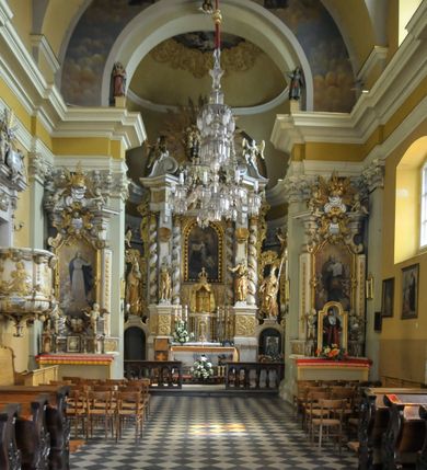 Zdjęcie nr 3:  Kościół usytuowany jest za północną pierzeją zabudowy rynku, w bezpośrednim sąsiedztwie świątyni parafialnej, po jej południowo-wschodniej stronie. 
Kościół orientowany, jest budowlą murowaną z cegły i kamienia (grubość murów od 70 do 160 cm), na rzucie podłużnym (24 m długość, 8,5 m szerokość). Korpus jednonawowy, trójprzęsłowy, prezbiterium jednoprzęsłowe, zamknięte półkolistą absydą, z dwukondygnacyjną przyległością od północy (zakrystia w przyziemiu). Nawę i prezbiterium przekrywa sklepienie kolebkowe z gurtami wspartymi na parach pilastrów dźwigających wydatne, obiegające wnętrze belkowanie. Nawę od prezbiterium oddziela półkolista arkada łuku tęczowego. Wnętrze doświetlają dwa rzędy okien zamkniętych łukami odcinkowymi. Artykulację ścian na zewnątrz stanowią płyciny ujmujące pary okien. Trójosiową fasadę artykułują pary osadzonych na cokole pilastrów – po dwie po bokach i jedna w polu środkowym wspierające belkowanie. W osi środkowej jest umiejscowiony ujęty profilowanym obramieniem otwór wejściowy z dwuskrzydłowymi drzwiami. Wyżej jest płytka, prostokątna wnęka, u góry zaś okno zamknięte łukiem odcinkowym. W osiach bocznych na cokole fasady osadzona para niszy z rzeźbami św. Piotra po lewej stronie i św. Pawła po prawej. Kapitele pilastrów zdobione motywami festonów owocowo-kwiatowych.   
Szczyt fasady ujęty parą spływów, zamknięty trójkątnie, po bokach flankowany parą cokołów zwieńczonych wazonami, pole środkowe wydzielone dwiema parami pilastrów zawiera półkoliście zamkniętą niszę z figurą Matki Boskiej, wyżej trójkąt Opatrzności Bożej ujęty promienistą glorią. Dachy korpusu i prezbiterium dwuspadowe, nad pierwszym przęsłem wieżyczka na sygnaturkę w formie latarni. Dach nad przyległością pulpitowy. Tynki kościoła w odcieniach bieli i szarości. 
