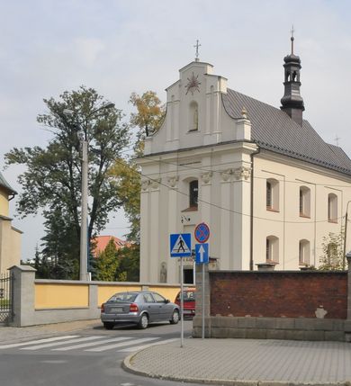 Zdjęcie nr 2:  Kościół usytuowany jest za północną pierzeją zabudowy rynku, w bezpośrednim sąsiedztwie świątyni parafialnej, po jej południowo-wschodniej stronie. 
Kościół orientowany, jest budowlą murowaną z cegły i kamienia (grubość murów od 70 do 160 cm), na rzucie podłużnym (24 m długość, 8,5 m szerokość). Korpus jednonawowy, trójprzęsłowy, prezbiterium jednoprzęsłowe, zamknięte półkolistą absydą, z dwukondygnacyjną przyległością od północy (zakrystia w przyziemiu). Nawę i prezbiterium przekrywa sklepienie kolebkowe z gurtami wspartymi na parach pilastrów dźwigających wydatne, obiegające wnętrze belkowanie. Nawę od prezbiterium oddziela półkolista arkada łuku tęczowego. Wnętrze doświetlają dwa rzędy okien zamkniętych łukami odcinkowymi. Artykulację ścian na zewnątrz stanowią płyciny ujmujące pary okien. Trójosiową fasadę artykułują pary osadzonych na cokole pilastrów – po dwie po bokach i jedna w polu środkowym wspierające belkowanie. W osi środkowej jest umiejscowiony ujęty profilowanym obramieniem otwór wejściowy z dwuskrzydłowymi drzwiami. Wyżej jest płytka, prostokątna wnęka, u góry zaś okno zamknięte łukiem odcinkowym. W osiach bocznych na cokole fasady osadzona para niszy z rzeźbami św. Piotra po lewej stronie i św. Pawła po prawej. Kapitele pilastrów zdobione motywami festonów owocowo-kwiatowych.   
Szczyt fasady ujęty parą spływów, zamknięty trójkątnie, po bokach flankowany parą cokołów zwieńczonych wazonami, pole środkowe wydzielone dwiema parami pilastrów zawiera półkoliście zamkniętą niszę z figurą Matki Boskiej, wyżej trójkąt Opatrzności Bożej ujęty promienistą glorią. Dachy korpusu i prezbiterium dwuspadowe, nad pierwszym przęsłem wieżyczka na sygnaturkę w formie latarni. Dach nad przyległością pulpitowy. Tynki kościoła w odcieniach bieli i szarości. 
