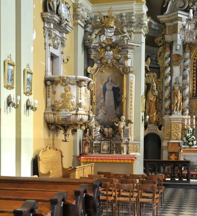 Zdjęcie nr 5:  Kościół usytuowany jest za północną pierzeją zabudowy rynku, w bezpośrednim sąsiedztwie świątyni parafialnej, po jej południowo-wschodniej stronie. 
Kościół orientowany, jest budowlą murowaną z cegły i kamienia (grubość murów od 70 do 160 cm), na rzucie podłużnym (24 m długość, 8,5 m szerokość). Korpus jednonawowy, trójprzęsłowy, prezbiterium jednoprzęsłowe, zamknięte półkolistą absydą, z dwukondygnacyjną przyległością od północy (zakrystia w przyziemiu). Nawę i prezbiterium przekrywa sklepienie kolebkowe z gurtami wspartymi na parach pilastrów dźwigających wydatne, obiegające wnętrze belkowanie. Nawę od prezbiterium oddziela półkolista arkada łuku tęczowego. Wnętrze doświetlają dwa rzędy okien zamkniętych łukami odcinkowymi. Artykulację ścian na zewnątrz stanowią płyciny ujmujące pary okien. Trójosiową fasadę artykułują pary osadzonych na cokole pilastrów – po dwie po bokach i jedna w polu środkowym wspierające belkowanie. W osi środkowej jest umiejscowiony ujęty profilowanym obramieniem otwór wejściowy z dwuskrzydłowymi drzwiami. Wyżej jest płytka, prostokątna wnęka, u góry zaś okno zamknięte łukiem odcinkowym. W osiach bocznych na cokole fasady osadzona para niszy z rzeźbami św. Piotra po lewej stronie i św. Pawła po prawej. Kapitele pilastrów zdobione motywami festonów owocowo-kwiatowych.   
Szczyt fasady ujęty parą spływów, zamknięty trójkątnie, po bokach flankowany parą cokołów zwieńczonych wazonami, pole środkowe wydzielone dwiema parami pilastrów zawiera półkoliście zamkniętą niszę z figurą Matki Boskiej, wyżej trójkąt Opatrzności Bożej ujęty promienistą glorią. Dachy korpusu i prezbiterium dwuspadowe, nad pierwszym przęsłem wieżyczka na sygnaturkę w formie latarni. Dach nad przyległością pulpitowy. Tynki kościoła w odcieniach bieli i szarości. 
