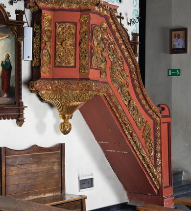 Zdjęcie nr 4: Ambona przyścienna dostępna jednotraktraktowymi schodami z pełną balustradą od strony wschodniej. Kosz na rzucie owalnym, opięty dostawionymi do boków pilastrami na rzucie trójkąta. W dolnej części złocony z podwieszoną szyszką. Podstawa walcowato wypukła dekorowana suchym akantem i jajownikiem. W polach kosza płyciny w kształcie stojącego prostokąta z symetryczną, złoconą dekoracją akantową. Trzony pilastrów wypełnione złoconym akantem o esowatym przebiegu wici. W zwieńczeniu wypukły, profilowany gzyms dekorowany palmetami I jajownikiem. Balustrada schodów zakończona pilastrem z prostokątną płyciną. W polu złocona dekoracją z suchego akantu. Struktura malowana w odcieniach ciemnej czerwieni i brązu, ornamentyka złocona.