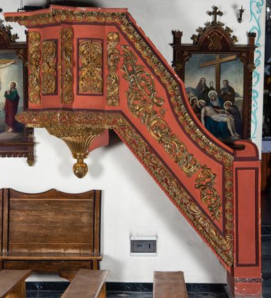 Zdjęcie nr 1: Ambona przyścienna dostępna jednotraktraktowymi schodami z pełną balustradą od strony wschodniej. Kosz na rzucie owalnym, opięty dostawionymi do boków pilastrami na rzucie trójkąta. W dolnej części złocony z podwieszoną szyszką. Podstawa walcowato wypukła dekorowana suchym akantem i jajownikiem. W polach kosza płyciny w kształcie stojącego prostokąta z symetryczną, złoconą dekoracją akantową. Trzony pilastrów wypełnione złoconym akantem o esowatym przebiegu wici. W zwieńczeniu wypukły, profilowany gzyms dekorowany palmetami I jajownikiem. Balustrada schodów zakończona pilastrem z prostokątną płyciną. W polu złocona dekoracją z suchego akantu. Struktura malowana w odcieniach ciemnej czerwieni i brązu, ornamentyka złocona.