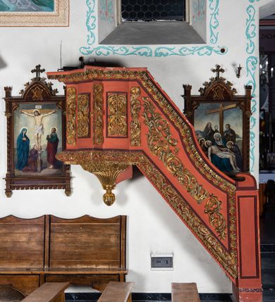 Zdjęcie nr 7: Ambona przyścienna dostępna jednotraktraktowymi schodami z pełną balustradą od strony wschodniej. Kosz na rzucie owalnym, opięty dostawionymi do boków pilastrami na rzucie trójkąta. W dolnej części złocony z podwieszoną szyszką. Podstawa walcowato wypukła dekorowana suchym akantem i jajownikiem. W polach kosza płyciny w kształcie stojącego prostokąta z symetryczną, złoconą dekoracją akantową. Trzony pilastrów wypełnione złoconym akantem o esowatym przebiegu wici. W zwieńczeniu wypukły, profilowany gzyms dekorowany palmetami I jajownikiem. Balustrada schodów zakończona pilastrem z prostokątną płyciną. W polu złocona dekoracją z suchego akantu. Struktura malowana w odcieniach ciemnej czerwieni i brązu, ornamentyka złocona.