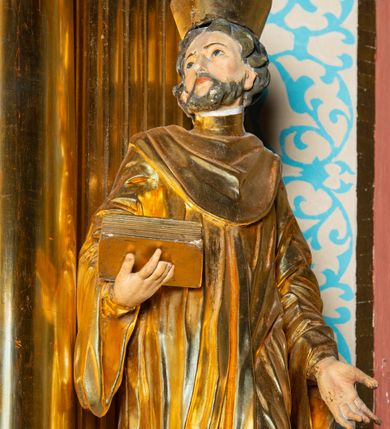 Zdjęcie nr 5: Rzeźba ścięta z tyłu, ustawiona na cokole. Święty ukazany frontalnie, w kontrapoście, z głową skierowaną w prawo. Lewą rękę ma opuszczonĄ w dół, prawą podtrzymuje zamkniętĄ księgę. Twarz ma kwadratową, o dużych, migdałowatych oczach, krótkim, prostym nosie i wąskich ustach, okolonĄ krótkimi, kręconymi włosami i brodą. Na głowie ma biret. Ubrany jest w togę profesorską o szerokich rękawach, z załogą (pelerynką). Polichromia ciała naturalistyczna, szaty i atrybuty złocone. 
