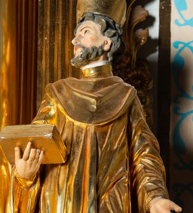 Zdjęcie nr 8: Rzeźba ścięta z tyłu, ustawiona na cokole. Święty ukazany frontalnie, w kontrapoście, z głową skierowaną w prawo. Lewą rękę ma opuszczonĄ w dół, prawą podtrzymuje zamkniętĄ księgę. Twarz ma kwadratową, o dużych, migdałowatych oczach, krótkim, prostym nosie i wąskich ustach, okolonĄ krótkimi, kręconymi włosami i brodą. Na głowie ma biret. Ubrany jest w togę profesorską o szerokich rękawach, z załogą (pelerynką). Polichromia ciała naturalistyczna, szaty i atrybuty złocone. 
