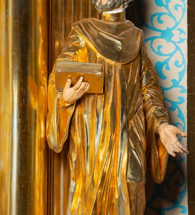 Zdjęcie nr 10: Rzeźba ścięta z tyłu, ustawiona na cokole. Święty ukazany frontalnie, w kontrapoście, z głową skierowaną w prawo. Lewą rękę ma opuszczonĄ w dół, prawą podtrzymuje zamkniętĄ księgę. Twarz ma kwadratową, o dużych, migdałowatych oczach, krótkim, prostym nosie i wąskich ustach, okolonĄ krótkimi, kręconymi włosami i brodą. Na głowie ma biret. Ubrany jest w togę profesorską o szerokich rękawach, z załogą (pelerynką). Polichromia ciała naturalistyczna, szaty i atrybuty złocone. 
