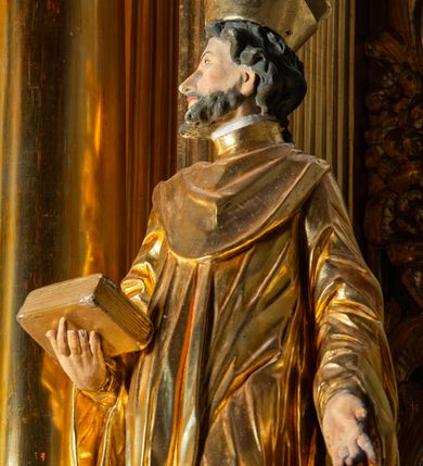 Zdjęcie nr 11: Rzeźba ścięta z tyłu, ustawiona na cokole. Święty ukazany frontalnie, w kontrapoście, z głową skierowaną w prawo. Lewą rękę ma opuszczonĄ w dół, prawą podtrzymuje zamkniętĄ księgę. Twarz ma kwadratową, o dużych, migdałowatych oczach, krótkim, prostym nosie i wąskich ustach, okolonĄ krótkimi, kręconymi włosami i brodą. Na głowie ma biret. Ubrany jest w togę profesorską o szerokich rękawach, z załogą (pelerynką). Polichromia ciała naturalistyczna, szaty i atrybuty złocone. 
