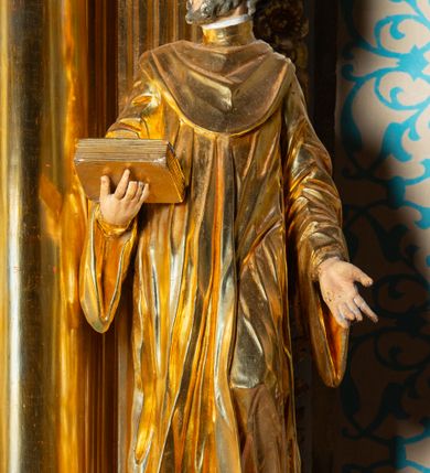 Zdjęcie nr 12: Rzeźba ścięta z tyłu, ustawiona na cokole. Święty ukazany frontalnie, w kontrapoście, z głową skierowaną w prawo. Lewą rękę ma opuszczonĄ w dół, prawą podtrzymuje zamkniętĄ księgę. Twarz ma kwadratową, o dużych, migdałowatych oczach, krótkim, prostym nosie i wąskich ustach, okolonĄ krótkimi, kręconymi włosami i brodą. Na głowie ma biret. Ubrany jest w togę profesorską o szerokich rękawach, z załogą (pelerynką). Polichromia ciała naturalistyczna, szaty i atrybuty złocone. 
