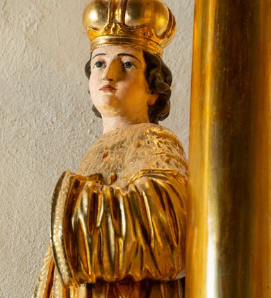 Zdjęcie nr 10: Rzeźba ścięta z tyłu, ustawiona na cokole. Święty ukazany frontalnie, w kontrapoście, z głową skierowaną w lewo. Prawą rękę ma opuszczoną, lewa spoczywa na piersi. Twarz ma kwadratową, o wyodrębnionej brodzie, młodzieńczych rysach, dużych, migdałowatych oczach, krótkim, prostym nosie i wąskich, rozchylonych ustach, okoloną krótkimi, kręconymi, brązowymi włosami. Na głowie ma mitrę książęcą. Ubrany jest w długą tunikę oraz rozchylony z przodu płaszcz królewski, o szerokich rękawach, z gronostajową pelerynką. Polichromia w partiach ciała naturalistyczna, szaty złocone. 

