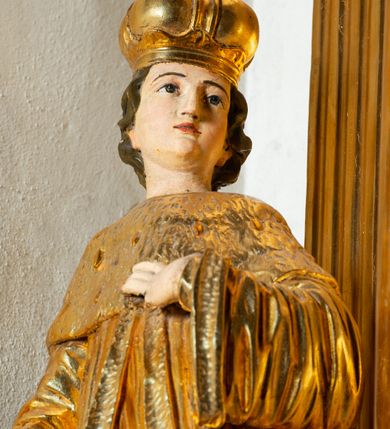 Zdjęcie nr 12: Rzeźba ścięta z tyłu, ustawiona na cokole. Święty ukazany frontalnie, w kontrapoście, z głową skierowaną w lewo. Prawą rękę ma opuszczoną, lewa spoczywa na piersi. Twarz ma kwadratową, o wyodrębnionej brodzie, młodzieńczych rysach, dużych, migdałowatych oczach, krótkim, prostym nosie i wąskich, rozchylonych ustach, okoloną krótkimi, kręconymi, brązowymi włosami. Na głowie ma mitrę książęcą. Ubrany jest w długą tunikę oraz rozchylony z przodu płaszcz królewski, o szerokich rękawach, z gronostajową pelerynką. Polichromia w partiach ciała naturalistyczna, szaty złocone. 

