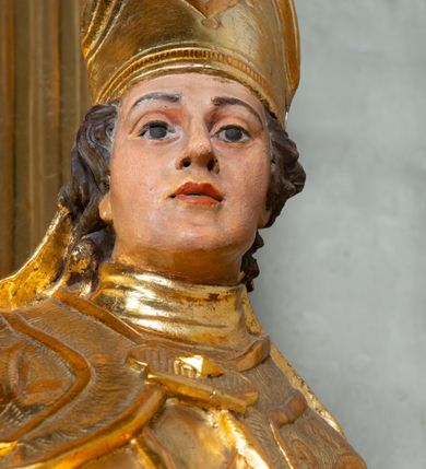 Zdjęcie nr 6: Rzeźba ścięta z tyłu, ustawiona na cokole. Święty ukazany frontalnie, w kontrapoście, z głową skierowaną w prawo.  Twarz ma kwadratową, z wyodrębnioną brodą, o dużych, migdałowatych oczach, krótkim, prostym nosie i wąskich ustach, okoloną krótkimi, brązowymi kręconymi włosami. Na głowie ma infułę z taśmami (fanones). Ubrany jest w albę, rokietę lamowaną frędzlą, kapę o rytym wzorze, zapiętą klamrą. Na piersi ma pektorał, a na dłoniach rękawiczki. Przy prawej nodze świętego półpostać Piotrowina, zwróconego w trzech czwartych w lewo, z uniesioną głową, o nagim torsie, z przerzuconym przez lewe ramię płaszczem. Polichromia w partiach ciał figur naturalistyczna, szaty złocone. 