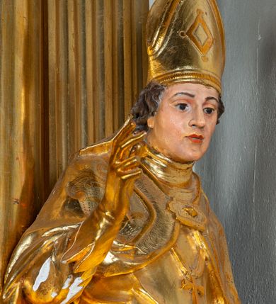 Zdjęcie nr 10: Rzeźba ścięta z tyłu, ustawiona na cokole. Święty ukazany frontalnie, w kontrapoście, z głową skierowaną w prawo.  Twarz ma kwadratową, z wyodrębnioną brodą, o dużych, migdałowatych oczach, krótkim, prostym nosie i wąskich ustach, okoloną krótkimi, brązowymi kręconymi włosami. Na głowie ma infułę z taśmami (fanones). Ubrany jest w albę, rokietę lamowaną frędzlą, kapę o rytym wzorze, zapiętą klamrą. Na piersi ma pektorał, a na dłoniach rękawiczki. Przy prawej nodze świętego półpostać Piotrowina, zwróconego w trzech czwartych w lewo, z uniesioną głową, o nagim torsie, z przerzuconym przez lewe ramię płaszczem. Polichromia w partiach ciał figur naturalistyczna, szaty złocone. 