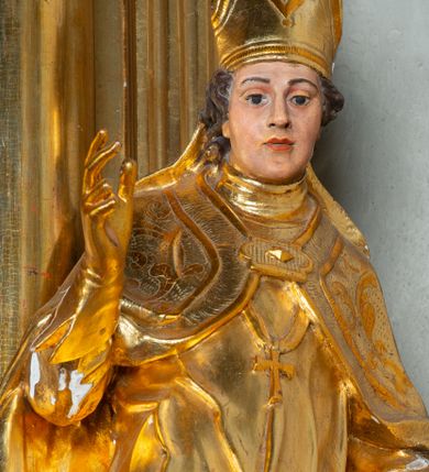 Zdjęcie nr 1: Rzeźba ścięta z tyłu, ustawiona na cokole. Święty ukazany frontalnie, w kontrapoście, z głową skierowaną w prawo.  Twarz ma kwadratową, z wyodrębnioną brodą, o dużych, migdałowatych oczach, krótkim, prostym nosie i wąskich ustach, okoloną krótkimi, brązowymi kręconymi włosami. Na głowie ma infułę z taśmami (fanones). Ubrany jest w albę, rokietę lamowaną frędzlą, kapę o rytym wzorze, zapiętą klamrą. Na piersi ma pektorał, a na dłoniach rękawiczki. Przy prawej nodze świętego półpostać Piotrowina, zwróconego w trzech czwartych w lewo, z uniesioną głową, o nagim torsie, z przerzuconym przez lewe ramię płaszczem. Polichromia w partiach ciał figur naturalistyczna, szaty złocone. 