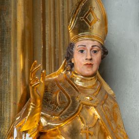 Zdjęcie nr 1: Rzeźba ścięta z tyłu, ustawiona na cokole. Święty ukazany frontalnie, w kontrapoście, z głową skierowaną w prawo.  Twarz ma kwadratową, z wyodrębnioną brodą, o dużych, migdałowatych oczach, krótkim, prostym nosie i wąskich ustach, okoloną krótkimi, brązowymi kręconymi włosami. Na głowie ma infułę z taśmami (fanones). Ubrany jest w albę, rokietę lamowaną frędzlą, kapę o rytym wzorze, zapiętą klamrą. Na piersi ma pektorał, a na dłoniach rękawiczki. Przy prawej nodze świętego półpostać Piotrowina, zwróconego w trzech czwartych w lewo, z uniesioną głową, o nagim torsie, z przerzuconym przez lewe ramię płaszczem. Polichromia w partiach ciał figur naturalistyczna, szaty złocone. 