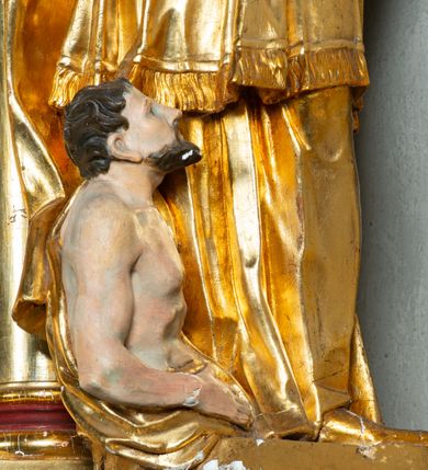 Zdjęcie nr 12: Rzeźba ścięta z tyłu, ustawiona na cokole. Święty ukazany frontalnie, w kontrapoście, z głową skierowaną w prawo.  Twarz ma kwadratową, z wyodrębnioną brodą, o dużych, migdałowatych oczach, krótkim, prostym nosie i wąskich ustach, okoloną krótkimi, brązowymi kręconymi włosami. Na głowie ma infułę z taśmami (fanones). Ubrany jest w albę, rokietę lamowaną frędzlą, kapę o rytym wzorze, zapiętą klamrą. Na piersi ma pektorał, a na dłoniach rękawiczki. Przy prawej nodze świętego półpostać Piotrowina, zwróconego w trzech czwartych w lewo, z uniesioną głową, o nagim torsie, z przerzuconym przez lewe ramię płaszczem. Polichromia w partiach ciał figur naturalistyczna, szaty złocone. 