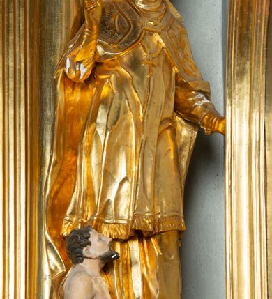 Zdjęcie nr 2: Rzeźba ścięta z tyłu, ustawiona na cokole. Święty ukazany frontalnie, w kontrapoście, z głową skierowaną w prawo.  Twarz ma kwadratową, z wyodrębnioną brodą, o dużych, migdałowatych oczach, krótkim, prostym nosie i wąskich ustach, okoloną krótkimi, brązowymi kręconymi włosami. Na głowie ma infułę z taśmami (fanones). Ubrany jest w albę, rokietę lamowaną frędzlą, kapę o rytym wzorze, zapiętą klamrą. Na piersi ma pektorał, a na dłoniach rękawiczki. Przy prawej nodze świętego półpostać Piotrowina, zwróconego w trzech czwartych w lewo, z uniesioną głową, o nagim torsie, z przerzuconym przez lewe ramię płaszczem. Polichromia w partiach ciał figur naturalistyczna, szaty złocone. 