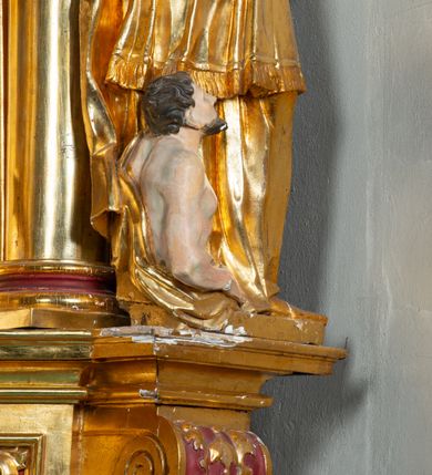 Zdjęcie nr 3: Rzeźba ścięta z tyłu, ustawiona na cokole. Święty ukazany frontalnie, w kontrapoście, z głową skierowaną w prawo.  Twarz ma kwadratową, z wyodrębnioną brodą, o dużych, migdałowatych oczach, krótkim, prostym nosie i wąskich ustach, okoloną krótkimi, brązowymi kręconymi włosami. Na głowie ma infułę z taśmami (fanones). Ubrany jest w albę, rokietę lamowaną frędzlą, kapę o rytym wzorze, zapiętą klamrą. Na piersi ma pektorał, a na dłoniach rękawiczki. Przy prawej nodze świętego półpostać Piotrowina, zwróconego w trzech czwartych w lewo, z uniesioną głową, o nagim torsie, z przerzuconym przez lewe ramię płaszczem. Polichromia w partiach ciał figur naturalistyczna, szaty złocone. 