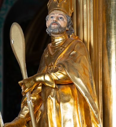 Zdjęcie nr 6: Rzeźba ścięta z tyłu, ustawiona na cokole. Święty ukazany frontalnie, w kontrapoście, z głową skierowaną w lewo. W prawej ręce trzyma laskę zakończoną potrójnym krzyżem (papieskim), w lewej krótkie wiosło. Twarz ma kwadratową, o dużych, migdałowatych oczach, krótkim, prostym nosie i małych, rozchylonych ustach, okoloną siwymi, kręconymi, krótkimi włosami i brodą. Na głowie ma infułę. Ubrany jest w albę, rokietę z dekoracyjną lamówką, kapę o rytym wzorze, na piersi ma paliusz. Polichromia ciała naturalistyczna, szaty i atrybuty złocone. 
