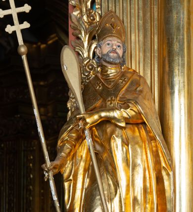 Zdjęcie nr 7: Rzeźba ścięta z tyłu, ustawiona na cokole. Święty ukazany frontalnie, w kontrapoście, z głową skierowaną w lewo. W prawej ręce trzyma laskę zakończoną potrójnym krzyżem (papieskim), w lewej krótkie wiosło. Twarz ma kwadratową, o dużych, migdałowatych oczach, krótkim, prostym nosie i małych, rozchylonych ustach, okoloną siwymi, kręconymi, krótkimi włosami i brodą. Na głowie ma infułę. Ubrany jest w albę, rokietę z dekoracyjną lamówką, kapę o rytym wzorze, na piersi ma paliusz. Polichromia ciała naturalistyczna, szaty i atrybuty złocone. 
