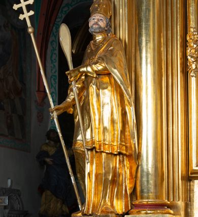 Zdjęcie nr 8: Rzeźba ścięta z tyłu, ustawiona na cokole. Święty ukazany frontalnie, w kontrapoście, z głową skierowaną w lewo. W prawej ręce trzyma laskę zakończoną potrójnym krzyżem (papieskim), w lewej krótkie wiosło. Twarz ma kwadratową, o dużych, migdałowatych oczach, krótkim, prostym nosie i małych, rozchylonych ustach, okoloną siwymi, kręconymi, krótkimi włosami i brodą. Na głowie ma infułę. Ubrany jest w albę, rokietę z dekoracyjną lamówką, kapę o rytym wzorze, na piersi ma paliusz. Polichromia ciała naturalistyczna, szaty i atrybuty złocone. 
