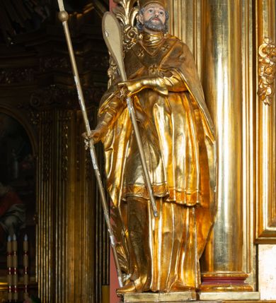 Zdjęcie nr 11: Rzeźba ścięta z tyłu, ustawiona na cokole. Święty ukazany frontalnie, w kontrapoście, z głową skierowaną w lewo. W prawej ręce trzyma laskę zakończoną potrójnym krzyżem (papieskim), w lewej krótkie wiosło. Twarz ma kwadratową, o dużych, migdałowatych oczach, krótkim, prostym nosie i małych, rozchylonych ustach, okoloną siwymi, kręconymi, krótkimi włosami i brodą. Na głowie ma infułę. Ubrany jest w albę, rokietę z dekoracyjną lamówką, kapę o rytym wzorze, na piersi ma paliusz. Polichromia ciała naturalistyczna, szaty i atrybuty złocone. 

