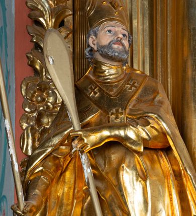 Zdjęcie nr 12: Rzeźba ścięta z tyłu, ustawiona na cokole. Święty ukazany frontalnie, w kontrapoście, z głową skierowaną w lewo. W prawej ręce trzyma laskę zakończoną potrójnym krzyżem (papieskim), w lewej krótkie wiosło. Twarz ma kwadratową, o dużych, migdałowatych oczach, krótkim, prostym nosie i małych, rozchylonych ustach, okoloną siwymi, kręconymi, krótkimi włosami i brodą. Na głowie ma infułę. Ubrany jest w albę, rokietę z dekoracyjną lamówką, kapę o rytym wzorze, na piersi ma paliusz. Polichromia ciała naturalistyczna, szaty i atrybuty złocone. 
