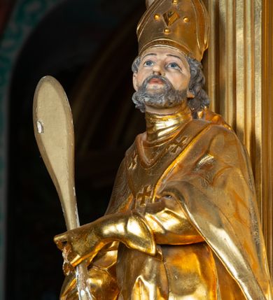 Zdjęcie nr 13: Rzeźba ścięta z tyłu, ustawiona na cokole. Święty ukazany frontalnie, w kontrapoście, z głową skierowaną w lewo. W prawej ręce trzyma laskę zakończoną potrójnym krzyżem (papieskim), w lewej krótkie wiosło. Twarz ma kwadratową, o dużych, migdałowatych oczach, krótkim, prostym nosie i małych, rozchylonych ustach, okoloną siwymi, kręconymi, krótkimi włosami i brodą. Na głowie ma infułę. Ubrany jest w albę, rokietę z dekoracyjną lamówką, kapę o rytym wzorze, na piersi ma paliusz. Polichromia ciała naturalistyczna, szaty i atrybuty złocone. 
