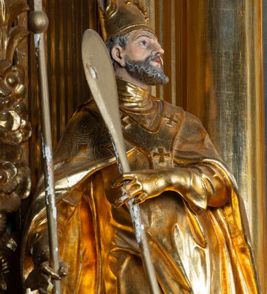 Zdjęcie nr 2: Rzeźba ścięta z tyłu, ustawiona na cokole. Święty ukazany frontalnie, w kontrapoście, z głową skierowaną w lewo. W prawej ręce trzyma laskę zakończoną potrójnym krzyżem (papieskim), w lewej krótkie wiosło. Twarz ma kwadratową, o dużych, migdałowatych oczach, krótkim, prostym nosie i małych, rozchylonych ustach, okoloną siwymi, kręconymi, krótkimi włosami i brodą. Na głowie ma infułę. Ubrany jest w albę, rokietę z dekoracyjną lamówką, kapę o rytym wzorze, na piersi ma paliusz. Polichromia ciała naturalistyczna, szaty i atrybuty złocone. 
