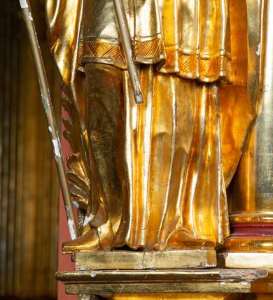 Zdjęcie nr 4: Rzeźba ścięta z tyłu, ustawiona na cokole. Święty ukazany frontalnie, w kontrapoście, z głową skierowaną w lewo. W prawej ręce trzyma laskę zakończoną potrójnym krzyżem (papieskim), w lewej krótkie wiosło. Twarz ma kwadratową, o dużych, migdałowatych oczach, krótkim, prostym nosie i małych, rozchylonych ustach, okoloną siwymi, kręconymi, krótkimi włosami i brodą. Na głowie ma infułę. Ubrany jest w albę, rokietę z dekoracyjną lamówką, kapę o rytym wzorze, na piersi ma paliusz. Polichromia ciała naturalistyczna, szaty i atrybuty złocone. 
