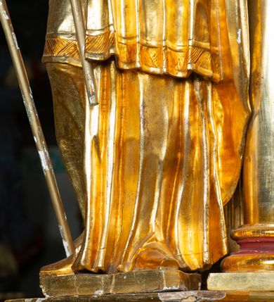 Zdjęcie nr 5: Rzeźba ścięta z tyłu, ustawiona na cokole. Święty ukazany frontalnie, w kontrapoście, z głową skierowaną w lewo. W prawej ręce trzyma laskę zakończoną potrójnym krzyżem (papieskim), w lewej krótkie wiosło. Twarz ma kwadratową, o dużych, migdałowatych oczach, krótkim, prostym nosie i małych, rozchylonych ustach, okoloną siwymi, kręconymi, krótkimi włosami i brodą. Na głowie ma infułę. Ubrany jest w albę, rokietę z dekoracyjną lamówką, kapę o rytym wzorze, na piersi ma paliusz. Polichromia ciała naturalistyczna, szaty i atrybuty złocone. 
