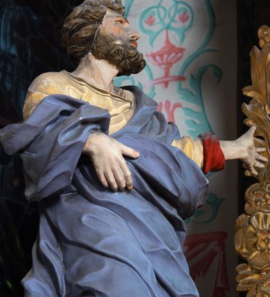 Zdjęcie nr 2: Rzeźba ścięta z tyłu i drążona. Święty zwrócony w trzech czwartych w lewo, w kontrapoście, z uniesiona głową. Lewą, uniesioną rękę wyciąga w bok, prawą podtrzymuje z przodu połę płaszcza. Twarz ma szczupłą, kwadratową, o migdałowatych oczach skierowanych w górę, ostrym profilu nosa oraz wąskich rozchylonych ustach. Włosy i broda brązowe, krótkie, rzeźbione w silnie skręcone, pojedyncze pukle. Ubrany jest w jasnożółtą, silnie drapowaną tunikę ze złoconą lamówką oraz niebieski płaszcz zarzucony na ramiona i zawinięty wokół ciała postaci, silnie skręcony, drapowany i rozwiany od spodu, zdobiony brązową lamówką i podbity czerwonym podbiciem. Polichromia ciała naturalistyczna.