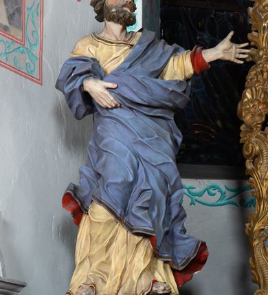 Zdjęcie nr 3: Rzeźba ścięta z tyłu i drążona. Święty zwrócony w trzech czwartych w lewo, w kontrapoście, z uniesiona głową. Lewą, uniesioną rękę wyciąga w bok, prawą podtrzymuje z przodu połę płaszcza. Twarz ma szczupłą, kwadratową, o migdałowatych oczach skierowanych w górę, ostrym profilu nosa oraz wąskich rozchylonych ustach. Włosy i broda brązowe, krótkie, rzeźbione w silnie skręcone, pojedyncze pukle. Ubrany jest w jasnożółtą, silnie drapowaną tunikę ze złoconą lamówką oraz niebieski płaszcz zarzucony na ramiona i zawinięty wokół ciała postaci, silnie skręcony, drapowany i rozwiany od spodu, zdobiony brązową lamówką i podbity czerwonym podbiciem. Polichromia ciała naturalistyczna.