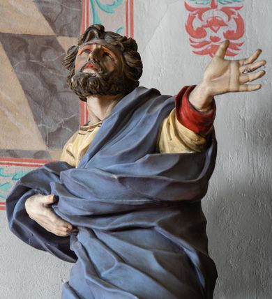 Zdjęcie nr 6: Rzeźba ścięta z tyłu i drążona. Święty zwrócony w trzech czwartych w lewo, w kontrapoście, z uniesiona głową. Lewą, uniesioną rękę wyciąga w bok, prawą podtrzymuje z przodu połę płaszcza. Twarz ma szczupłą, kwadratową, o migdałowatych oczach skierowanych w górę, ostrym profilu nosa oraz wąskich rozchylonych ustach. Włosy i broda brązowe, krótkie, rzeźbione w silnie skręcone, pojedyncze pukle. Ubrany jest w jasnożółtą, silnie drapowaną tunikę ze złoconą lamówką oraz niebieski płaszcz zarzucony na ramiona i zawinięty wokół ciała postaci, silnie skręcony, drapowany i rozwiany od spodu, zdobiony brązową lamówką i podbity czerwonym podbiciem. Polichromia ciała naturalistyczna.