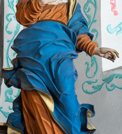 Zdjęcie nr 3: Rzeźba ścięta z tyłu i drążona. Święta zwrócona w trzech czwartych w prawo, w kontrapoście, na bosych stopach. Prawą, ugiętą rękę unosi na tle piersi, lewą wyciąga w bok. Twarz ma szczupłą, o migdałowatych oczach skierowanych w górę, ostrym profilu nosa i wąskich ustach, okoloną brązowymi, falowanymi włosami, ukrytymi pod welonem. Ubrana jest w jasnoróżową, silnie drapowaną suknię przepasaną w biodrze oraz niebieski płaszcz z czerwoną lamówką zawinięty wokół lewego ramienia i ciała świętej, silnie drapowany, ekspresyjnie rozwiany od dołu. Na głowie ma biały welon, odsłaniający profil twarzy, zawinięty wokół u szyi. Polichromia ciała naturalistyczna.