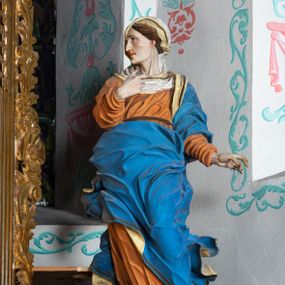 Zdjęcie nr 1: Rzeźba ścięta z tyłu i drążona. Święta zwrócona w trzech czwartych w prawo, w kontrapoście, na bosych stopach. Prawą, ugiętą rękę unosi na tle piersi, lewą wyciąga w bok. Twarz ma szczupłą, o migdałowatych oczach skierowanych w górę, ostrym profilu nosa i wąskich ustach, okoloną brązowymi, falowanymi włosami, ukrytymi pod welonem. Ubrana jest w jasnoróżową, silnie drapowaną suknię przepasaną w biodrze oraz niebieski płaszcz z czerwoną lamówką zawinięty wokół lewego ramienia i ciała świętej, silnie drapowany, ekspresyjnie rozwiany od dołu. Na głowie ma biały welon, odsłaniający profil twarzy, zawinięty wokół u szyi. Polichromia ciała naturalistyczna.