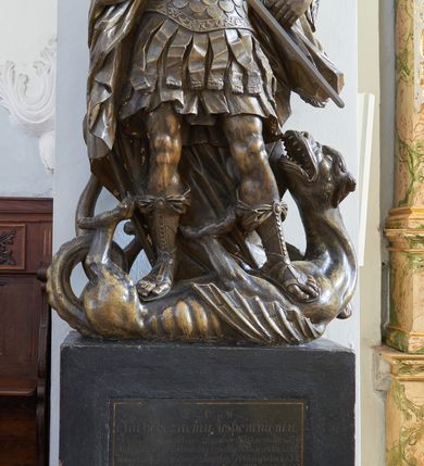 Zdjęcie nr 2: Rzeźba i nagrobek przyścienne. Nagrobek prostopadłościenny na cokole. Na nagrobku zamieszczona inskrypcja na marmurowej, wprawionej tablicy o wymiarach 70x 58 cm: „D.O.M./ Ku pobożnemu wspomnieniu/ Józefa Piątkowskiego zmarłego 1829 r: w wieku lat 70./ Magdaleny Piątkowskiej zmarłej 1855 r: w wieku lat 89./ Kaspra Piątkowskiego zmarłego 1847 r: w wieku lat 52./ Antoniego Piątkowskiego zmarłego 1847 r: w wieku lat 47./ Maryanny z Piątkowskich Łozińskiej/ zmarłej w 1849 r: w wieku lat 50./ Wiktoryj Nowakowskiej w Zakonie Franciszki zmarłej w 1854 r: w wieku lat 19/X: Piątkowski Serafin/ ten pomnik postawił”. Rzeźba ustawiona na nagrobku, przedstawia Michała Archanioła stojącego na smoku. Budowa ciała muskularna.Anioł w dynamicznej pozie, kontrapoście.Ciężar ciała oparty na prawej nodze. Tors i głowa silnie przechylone w lewą stronę. Ręce uniesione do wysokości ramion, ugięte w łokciach, przedramiona skierowane w dół. U ramion duże, dynamicznie ukształtowane skrzydła. Włosy kręcone, długością sięgające ramion, twarz o młodzieńczych rysach.W prawej dłoni trzyma miecz, skierowany ukośnie w dół, w stronę głowy smoka. Ubrany w zbroję łuskową, w dolnej partii zakończoną fartuchem złożonym zpasów, hełm z obfitym pióropuszem oraz sandały sięgające połowy łydki. Postać odziana jest w dekoracyjnie kształtowany płaszcz. Smok leżący na grzbiecie, pod stopami Archanioła, ma głowę i łapy wyciągnięte ku górze. Na grzbiecie łuska oraz skrzydła.