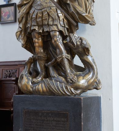 Zdjęcie nr 1: Rzeźba i nagrobek przyścienne. Nagrobek prostopadłościenny na cokole. Na nagrobku zamieszczona inskrypcja na marmurowej, wprawionej tablicy o wymiarach 70x 58 cm: „D.O.M./ Ku pobożnemu wspomnieniu/ Józefa Piątkowskiego zmarłego 1829 r: w wieku lat 70./ Magdaleny Piątkowskiej zmarłej 1855 r: w wieku lat 89./ Kaspra Piątkowskiego zmarłego 1847 r: w wieku lat 52./ Antoniego Piątkowskiego zmarłego 1847 r: w wieku lat 47./ Maryanny z Piątkowskich Łozińskiej/ zmarłej w 1849 r: w wieku lat 50./ Wiktoryj Nowakowskiej w Zakonie Franciszki zmarłej w 1854 r: w wieku lat 19/X: Piątkowski Serafin/ ten pomnik postawił”. Rzeźba ustawiona na nagrobku, przedstawia Michała Archanioła stojącego na smoku. Budowa ciała muskularna.Anioł w dynamicznej pozie, kontrapoście.Ciężar ciała oparty na prawej nodze. Tors i głowa silnie przechylone w lewą stronę. Ręce uniesione do wysokości ramion, ugięte w łokciach, przedramiona skierowane w dół. U ramion duże, dynamicznie ukształtowane skrzydła. Włosy kręcone, długością sięgające ramion, twarz o młodzieńczych rysach.W prawej dłoni trzyma miecz, skierowany ukośnie w dół, w stronę głowy smoka. Ubrany w zbroję łuskową, w dolnej partii zakończoną fartuchem złożonym zpasów, hełm z obfitym pióropuszem oraz sandały sięgające połowy łydki. Postać odziana jest w dekoracyjnie kształtowany płaszcz. Smok leżący na grzbiecie, pod stopami Archanioła, ma głowę i łapy wyciągnięte ku górze. Na grzbiecie łuska oraz skrzydła.