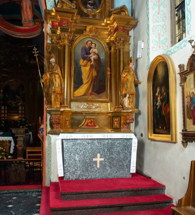 Zdjęcie nr 34: Nastawa architektoniczna, jednoosiowa, dwukondygnacyjna. Ołtarz prostopadłościenny, zwężający się ku dołowi, murowany, obłożony kamiennymi płytami, z krzyżem na froncie. 
Nastawa na niskim cokole, z wyodrębnionymi dwoma, ukośnie ustawionymi prostopadłościanami, z konsolami, dekorowanymi akantem i kampanulami. Pośrodku cokołu wyodrębniony postument dekorowany płyciną z akantem. Kondygnacja flankowana dwiema parami pilastrów o kanelowanych trzonach oraz ustawioną przed nimi parą kolumn o kapitelach kompozytowych. Podpory dźwigają pełne belkowanie, wyłamane na ich osi, z silnie wysuniętym gzymsem, z przerwanym architrawem i fryzem nad polem środkowym. Na belkowaniu fragmenty przerwanego, wolutowego przyczółka, zwrócone pod kątem, do wewnątrz. W polu środkowym wnęka w formie stojącego prostokąta zamkniętego łukiem półkolistym, ujęta profilowaną ramą dekorowaną na bokach kwiatami, zwieńczoną muszlą. We wnęce obraz św. Józefa z Dzieciątkiem. Poniżej wnęki płaskorzeźbione narzędzia stolarskie (piła, siekiera, kątownik, obcęgi) i stylizowany monogram świętego. Na konsolach, po bokach nastawy ustawione figury św. Wojciecha i św. Stanisława. Retabulum flankowane uszami utworzonymi z karbowanej wstęgi i ornamentu roślinnego. Druga kondygnacja węższa, ujęta ustawionymi pod kątem pilastrami, o trzonach dekorowanych kampanulami. Pilastry podtrzymują gzyms wyłamany na ich osi, nad polem przyjmujący kształt łuku półkolistego nadwieszonego. Na nim ustawione trzy wazony z kwiatami. W polu obraz św. Jana Kantego w formie stojącego owalu, ujęty ramą, dekorowaną ornamentem roślinnym. Struktura niemal w całości złocona, elementy cokołu i fryzu polichromowane w kolorze czerwonym, ornamentyka złocona. 
