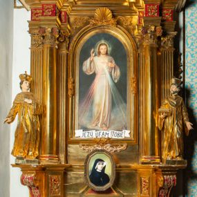 Zdjęcie nr 1: Nastawa architektoniczna, jednoosiowa, dwukondygnacyjna. Ołtarz murowany, prostopadłościenny, obity metalową blachą, zdobioną trybowaną kratownicą i gwiazdkami. Wewnątrz grób Chrystusa ze współczesną rzeźbą. Nastawa na niskim cokole, z wyodrębnionymi dwoma, ukośnie ustawionymi prostopadłościanami, z konsolami, dekorowanymi akantem i kampanulami. Pośrodku cokołu zamontowany obraz św. Faustyny w kształcie wieloboku. Kondygnacja flankowana dwiema parami pilastrów o kanelowanych trzonach oraz ustawioną przed nimi parą kolumn o kapitelach kompozytowych. Podpory dźwigają pełne belkowanie, wyłamane na ich osi, z silnie wysuniętym gzymsem, z przerwanym architrawem i fryzem nad polem środkowym. Na belkowaniu fragmenty przerwanego, wolutowego przyczółka, zwrócone pod kątem, do wewnątrz. W polu środkowym wnęka w formie stojącego prostokąta zamkniętego łukiem półkolistym, ujęta profilowaną ramą dekorowaną na bokach kwiatami, zwieńczoną muszlą. We wnęce obraz „Jezu Ufam Tobie”. Na konsolach, po bokach nastawy ustawione figury św. Kazimierza i św. Jana Kantego. Nastawa flankowana uszami utworzonymi z karbowanej wstęgi i ornamentu roślinnego. Druga kondygnacja węższa, flankowana ustawionymi pod kątem pilastrami, o trzonach dekorowanych kampanulami. Pilastry podtrzymują gzyms wyłamany na ich osi, nad polem przyjmujący kształt łuku półkolistego nadwieszonego. Na nim ustawione dwa wazony z kwiatami i bukiet kwiatów. W polu obraz św. Joachima w formie stojącego owalu, ujęty ramą, dekorowaną ornamentem roślinnym. Struktura niemal w całości złocona, elementy cokołu i fryzu polichromowane w kolorze czerwonym, ornamentyka złocona. 


