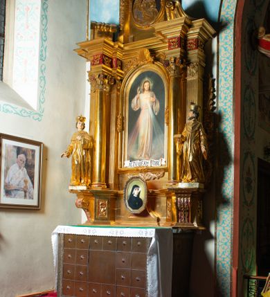 Zdjęcie nr 12: Nastawa architektoniczna, jednoosiowa, dwukondygnacyjna. Ołtarz murowany, prostopadłościenny, obity metalową blachą, zdobioną trybowaną kratownicą i gwiazdkami. Wewnątrz grób Chrystusa ze współczesną rzeźbą. Nastawa na niskim cokole, z wyodrębnionymi dwoma, ukośnie ustawionymi prostopadłościanami, z konsolami, dekorowanymi akantem i kampanulami. Pośrodku cokołu zamontowany obraz św. Faustyny w kształcie wieloboku. Kondygnacja flankowana dwiema parami pilastrów o kanelowanych trzonach oraz ustawioną przed nimi parą kolumn o kapitelach kompozytowych. Podpory dźwigają pełne belkowanie, wyłamane na ich osi, z silnie wysuniętym gzymsem, z przerwanym architrawem i fryzem nad polem środkowym. Na belkowaniu fragmenty przerwanego, wolutowego przyczółka, zwrócone pod kątem, do wewnątrz. W polu środkowym wnęka w formie stojącego prostokąta zamkniętego łukiem półkolistym, ujęta profilowaną ramą dekorowaną na bokach kwiatami, zwieńczoną muszlą. We wnęce obraz „Jezu Ufam Tobie”. Na konsolach, po bokach nastawy ustawione figury św. Kazimierza i św. Jana Kantego. Nastawa flankowana uszami utworzonymi z karbowanej wstęgi i ornamentu roślinnego. Druga kondygnacja węższa, flankowana ustawionymi pod kątem pilastrami, o trzonach dekorowanych kampanulami. Pilastry podtrzymują gzyms wyłamany na ich osi, nad polem przyjmujący kształt łuku półkolistego nadwieszonego. Na nim ustawione dwa wazony z kwiatami i bukiet kwiatów. W polu obraz św. Joachima w formie stojącego owalu, ujęty ramą, dekorowaną ornamentem roślinnym. Struktura niemal w całości złocona, elementy cokołu i fryzu polichromowane w kolorze czerwonym, ornamentyka złocona. 


