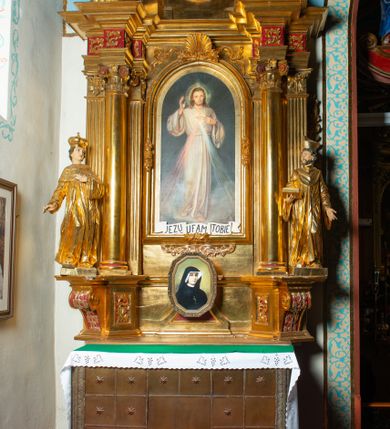 Zdjęcie nr 23: Nastawa architektoniczna, jednoosiowa, dwukondygnacyjna. Ołtarz murowany, prostopadłościenny, obity metalową blachą, zdobioną trybowaną kratownicą i gwiazdkami. Wewnątrz grób Chrystusa ze współczesną rzeźbą. Nastawa na niskim cokole, z wyodrębnionymi dwoma, ukośnie ustawionymi prostopadłościanami, z konsolami, dekorowanymi akantem i kampanulami. Pośrodku cokołu zamontowany obraz św. Faustyny w kształcie wieloboku. Kondygnacja flankowana dwiema parami pilastrów o kanelowanych trzonach oraz ustawioną przed nimi parą kolumn o kapitelach kompozytowych. Podpory dźwigają pełne belkowanie, wyłamane na ich osi, z silnie wysuniętym gzymsem, z przerwanym architrawem i fryzem nad polem środkowym. Na belkowaniu fragmenty przerwanego, wolutowego przyczółka, zwrócone pod kątem, do wewnątrz. W polu środkowym wnęka w formie stojącego prostokąta zamkniętego łukiem półkolistym, ujęta profilowaną ramą dekorowaną na bokach kwiatami, zwieńczoną muszlą. We wnęce obraz „Jezu Ufam Tobie”. Na konsolach, po bokach nastawy ustawione figury św. Kazimierza i św. Jana Kantego. Nastawa flankowana uszami utworzonymi z karbowanej wstęgi i ornamentu roślinnego. Druga kondygnacja węższa, flankowana ustawionymi pod kątem pilastrami, o trzonach dekorowanych kampanulami. Pilastry podtrzymują gzyms wyłamany na ich osi, nad polem przyjmujący kształt łuku półkolistego nadwieszonego. Na nim ustawione dwa wazony z kwiatami i bukiet kwiatów. W polu obraz św. Joachima w formie stojącego owalu, ujęty ramą, dekorowaną ornamentem roślinnym. Struktura niemal w całości złocona, elementy cokołu i fryzu polichromowane w kolorze czerwonym, ornamentyka złocona. 


